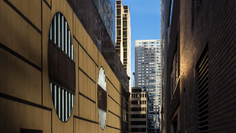 A narrow space between two buildings, with another skyscraper in the distance. The sky is very blue. The light reflecting off the buildings is golden.