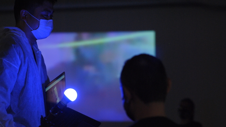 On the left, a figure wearing non-surgical mask and is holding a lightbulb and a computer tablet. On the right, an audience member with short hair, viewed from behind. In the background an indistinct projection on the wall.