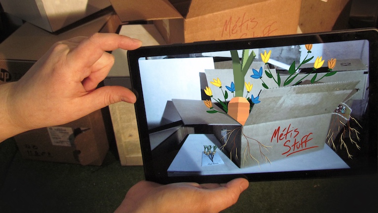 A person's hands holding a computer tablet in front of a group of cardboard boxes. Pictured on the tablet is a cardboard box labelled "Métis Stuff". Emerging from the box are drawings of flowers, carrots and roots.