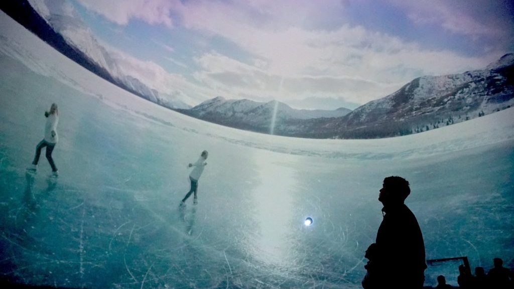 A dark shadow of a person watches SESQUI's 360 video of skaters on ice.