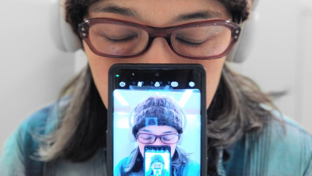 Show Image for SmartSmart - Adrienne wears a a brown beanie, red glasses, a blue shirt, and white headphones. She is holding up a smart phone. The screen is the same image of Adirenne but smaller. This repeats and gets smaller until we can no longer see the image.